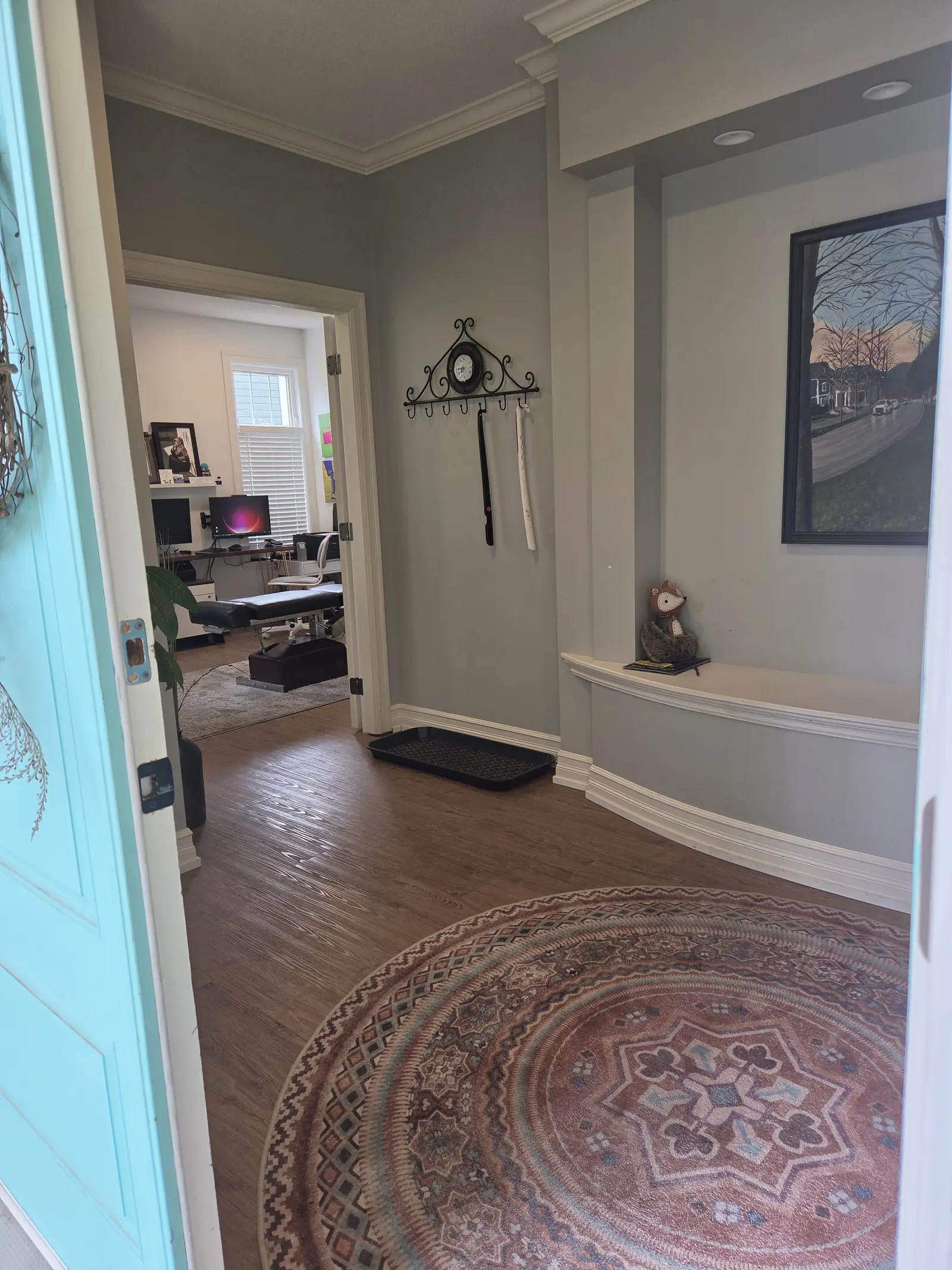 The entryway seen from the open front door: a round rug, a bench nook under a painting, and the treatment room beyond